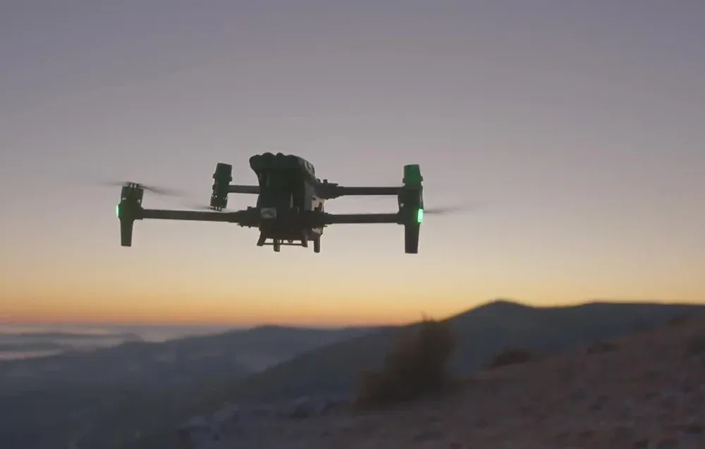 Dron DJI Matrice 30T lecący na tle zachodzącego słońca nad górzystym terenem.