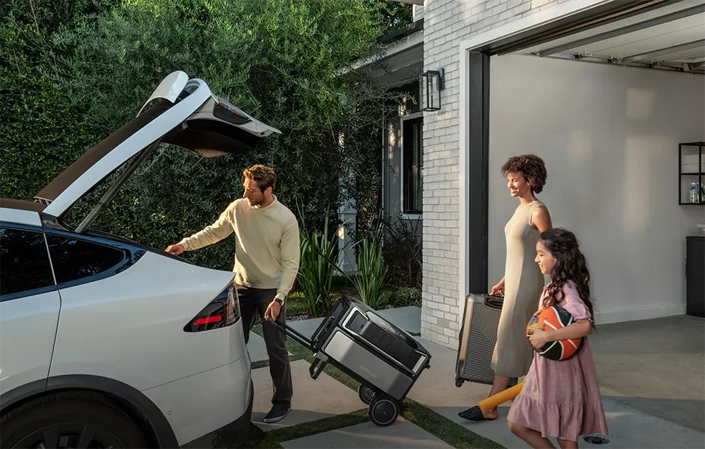 Family loading a portable power station into the trunk of a white car in front of a garage