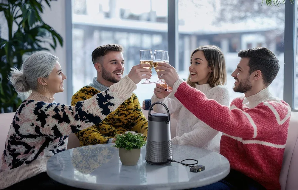 Czworo przyjaciół wznosi toast kieliszkami przy stole z głośnikiem i adapterem Bluetooth w jasnym pokoju