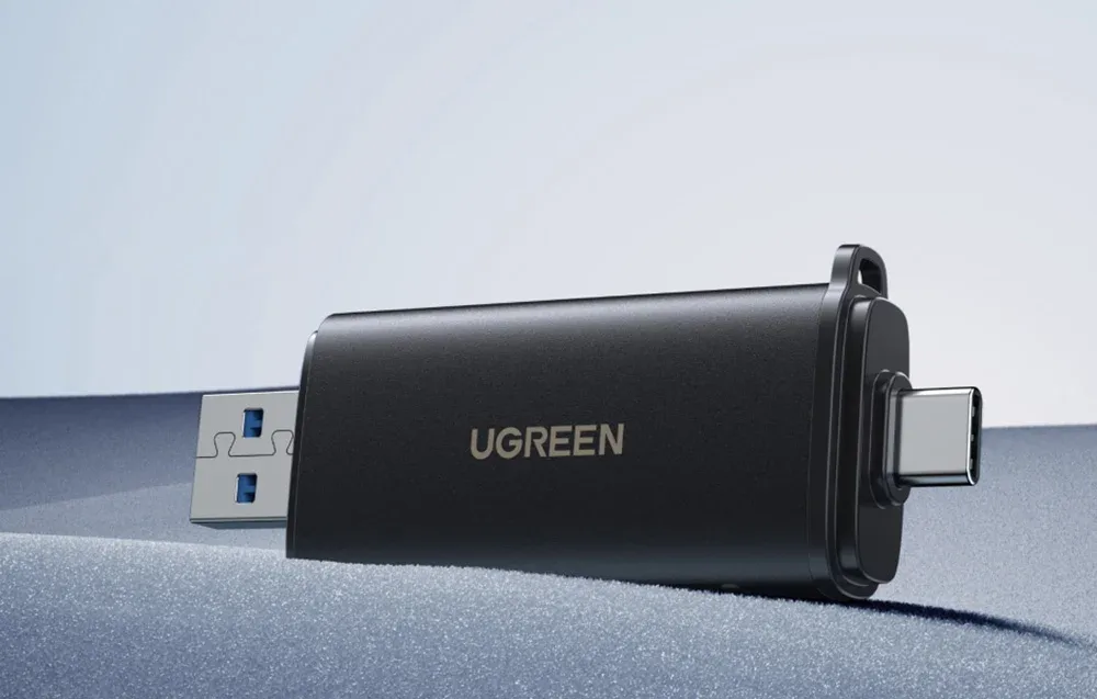 Czarny adapter UGREEN z wtyczkami USB-A i USB-C leży na szarej powierzchni na minimalistycznym tle