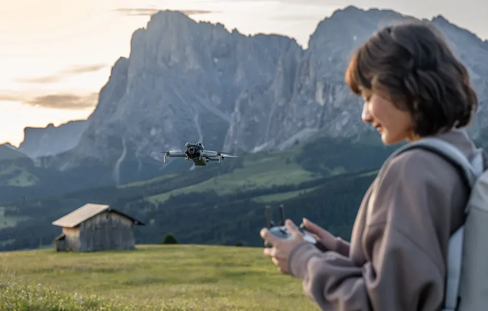 Kobieta steruje dronem na tle górskiego krajobrazu z drewnianą chatą na łące