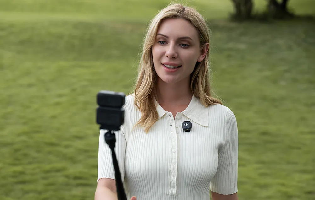 Kobieta na tle trawnika mówi do kamery na statywie, mając mikrofon DJI przypięty do koszulki
