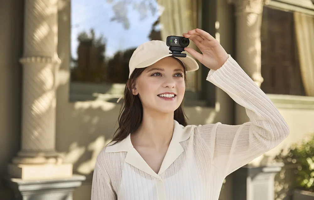 Kobieta w beżowej czapce z przypiętą na daszku kamerą DJI Osmo Action Nano patrzy w dal i uśmiecha się