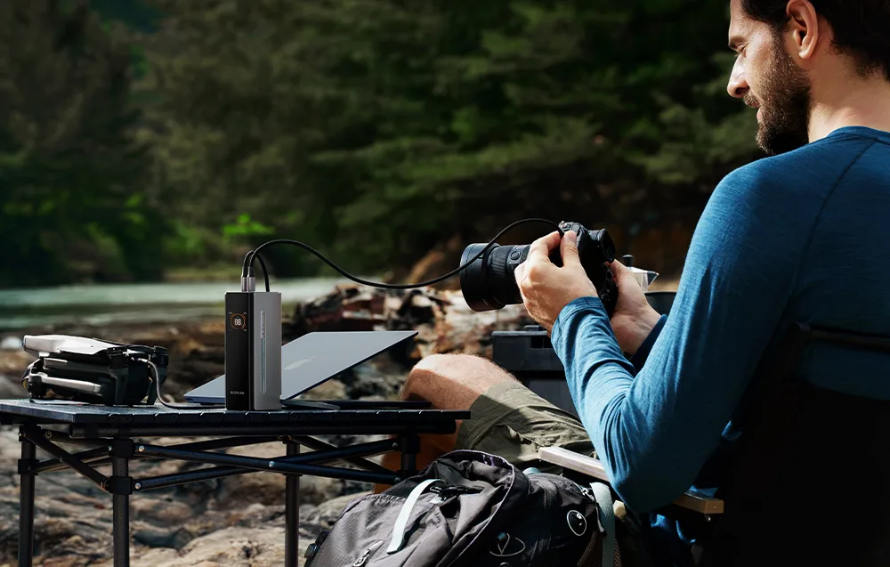 Man holding a camera while sitting near a table with a laptop, drone, and EcoFlow power bank outdoors.