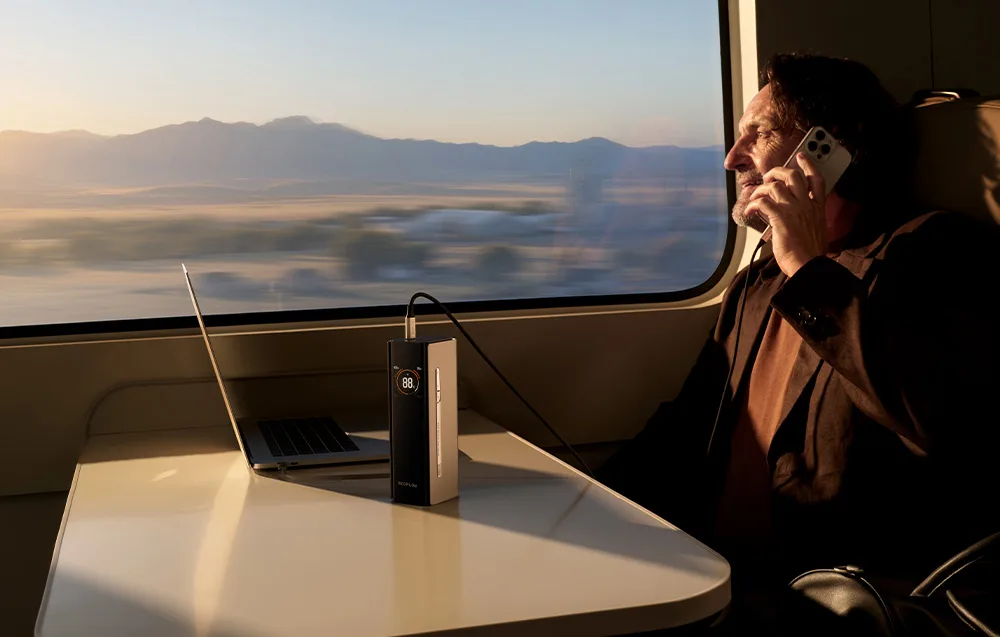 Man sitting on a train talking on the phone next to a laptop connected to the EcoFlow power bank with display.
