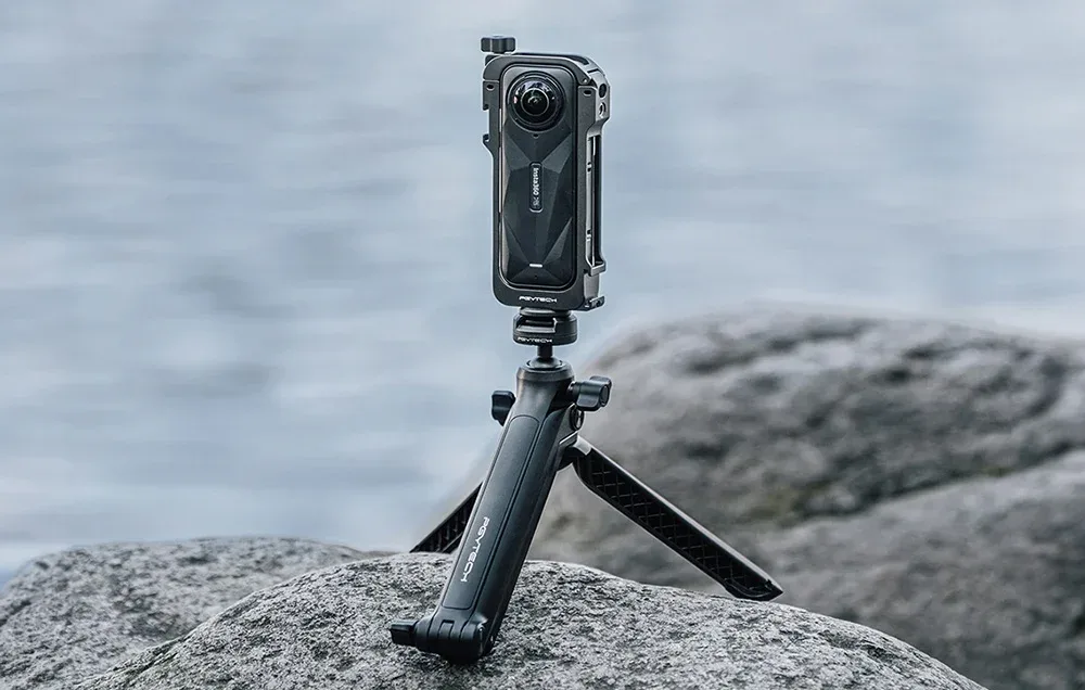 Kamera insta360 X5 w obudowie i na statywie leży na kamieniu. W tle widać wodę.