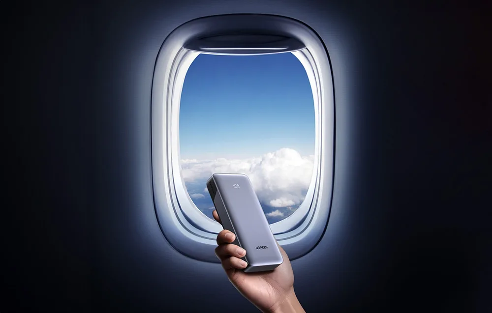 Hand holding a Ugreen power bank near an airplane window with a view of the sky
