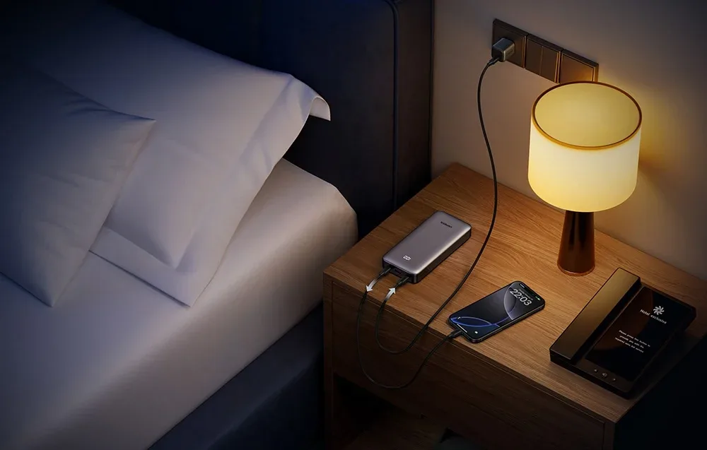 Ugreen power bank charging a phone on a bedside table next to a bed and lamp