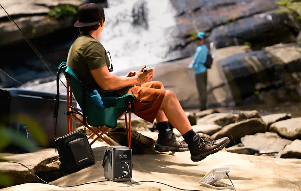 Person sitting on a camping chair near a waterfall with the EcoFlow TRAIL 300 DC portable power station on the ground