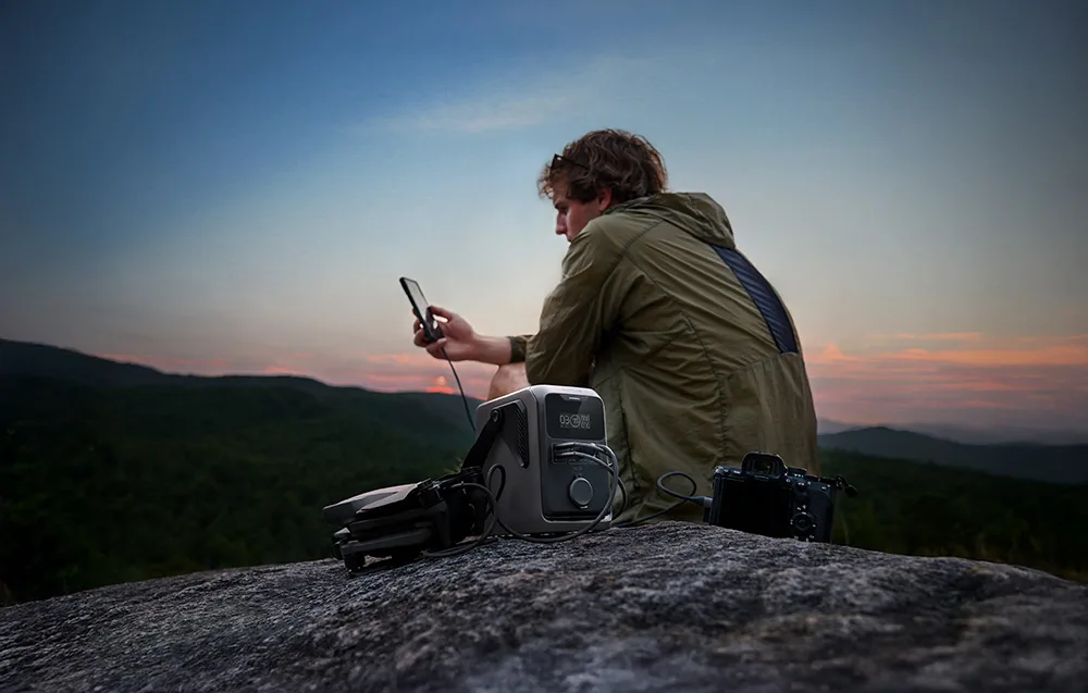 Man using a smartphone at sunset next to the EcoFlow TRAIL 300 DC with a drone and camera on a rock