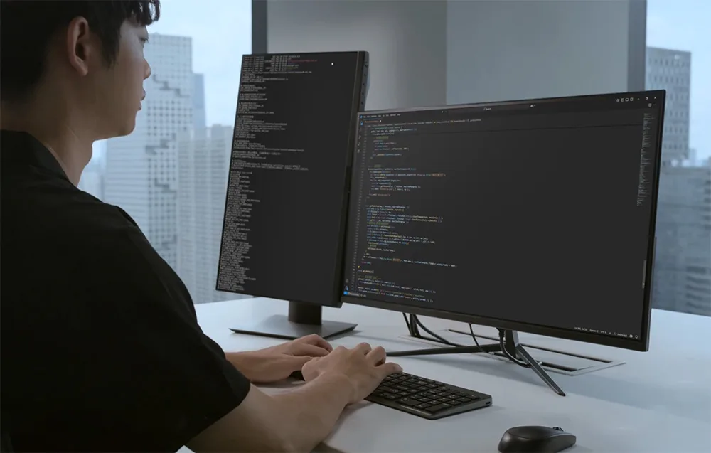 Programista przed dwoma monitorami wyświetlającymi kod źródłowy, ręce na klawiaturze