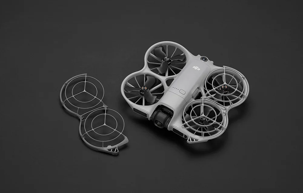 Szary dron DJI z czterema śmigłami, z czego dwa są osłonięte zdejmowaną osłoną widoczną obok na czarnym tle