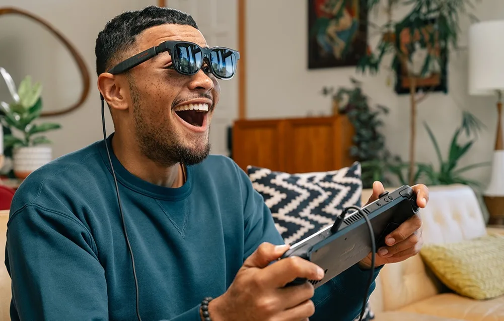 Man wearing VITURE XR Luma Pro glasses laughing while playing video games with a controller in the living room