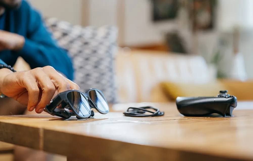 Hand reaching for black VITURE XR Luma Pro glasses on a wooden table next to a cable and game controller