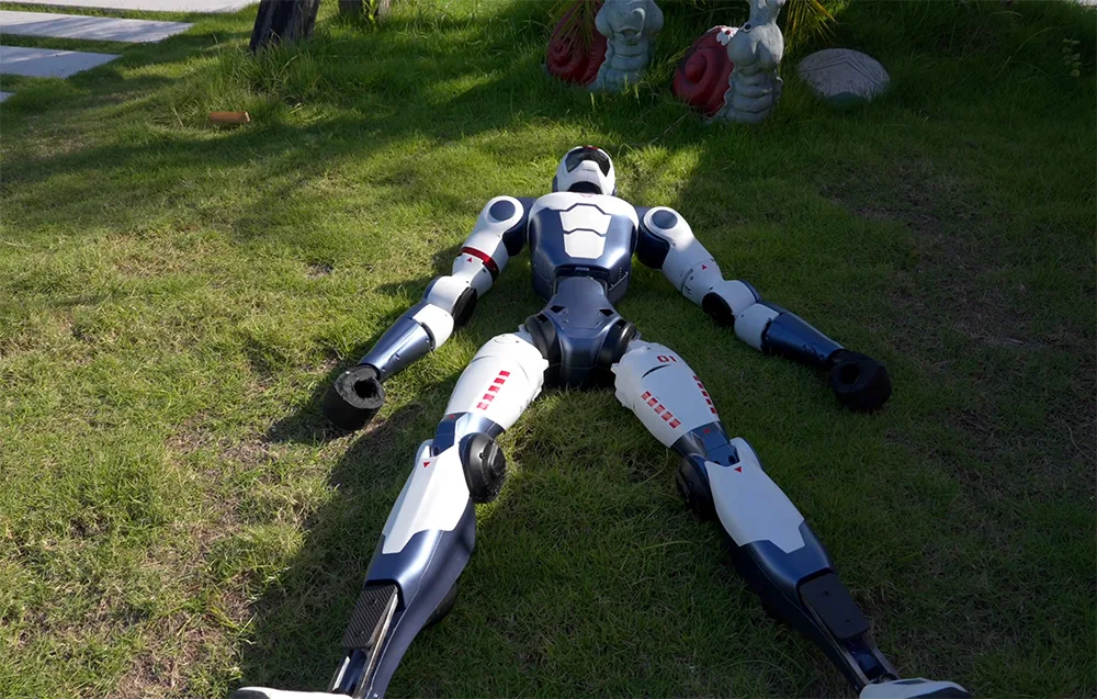 UNITREE R1 Basic humanoid robot lying on grass in the shade with arms and legs spread out