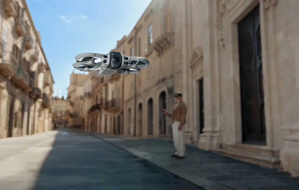 Сріблястий дрон DJI Avata завис у вузькій історичній вуличці, на задньому плані чоловік у FPV-окулярах
