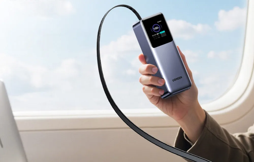 Hand holding Ugreen PB726 power bank with display visible near an airplane window