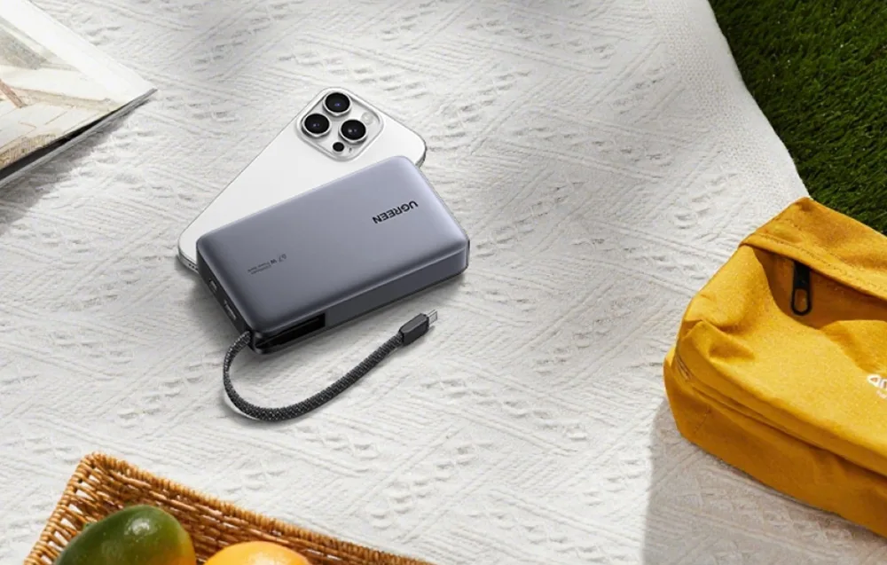 Ugreen PB550 power bank and iPhone on a picnic blanket next to a yellow bag, showing outdoor and travel use.