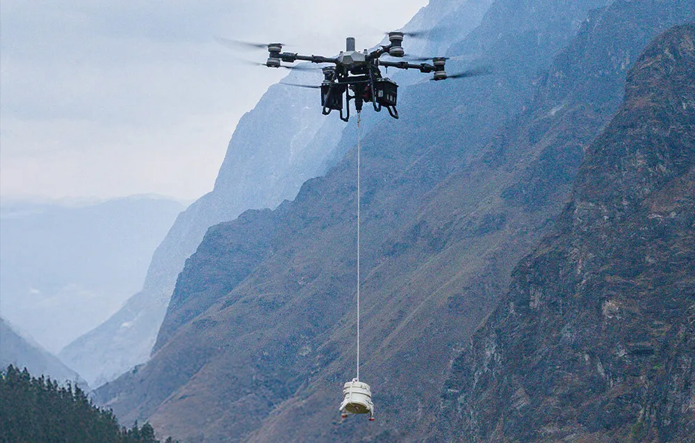 Dron transportowy lecący w górach, przenoszący na długiej lince biały, cylindryczny ładunek