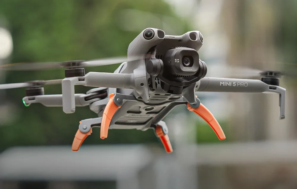 Latający dron DJI Mini 5 Pro z rozłożonymi pomarańczowymi nogami, unoszący się w powietrzu