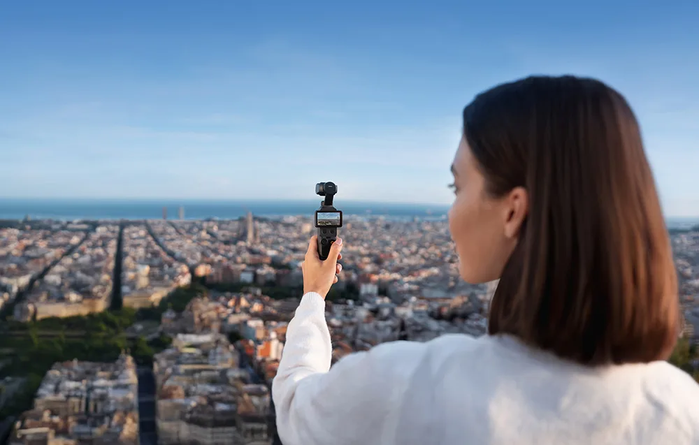 Widok zza pleców kobiety patrzącej na panoramę miasta, która trzyma w dłoni kamerę DJI Osmo Pocket 4 z podglądem budynków na ekranie