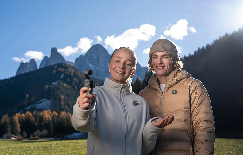 Uśmiechnięta kobieta w jasnym polarze trzyma kamerę DJI Osmo Pocket 4, pozując do nagrania z mężczyzną w brązowej kurtce na tle ośnieżonych szczytów górskich