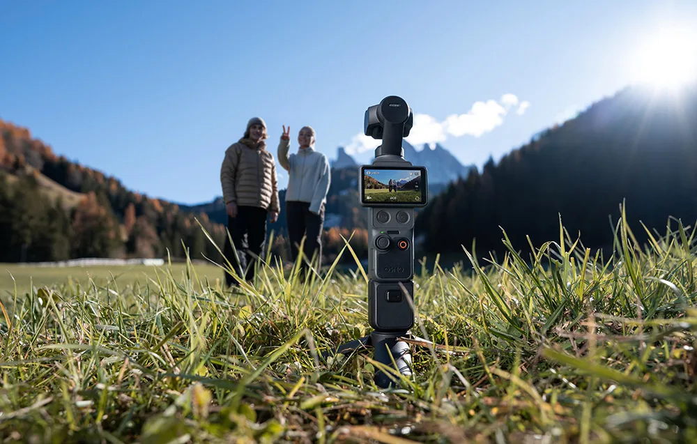 Kamera DJI Osmo Pocket 4 stoi na statywie w trawie i nagrywa dwójkę ludzi stojących w oddali na tle górskiej doliny