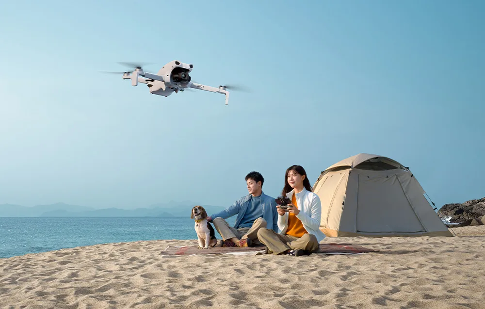Woman and man relaxing on a beach beside a tent while a white drone hovers above, controlled by the woman with a remote controller