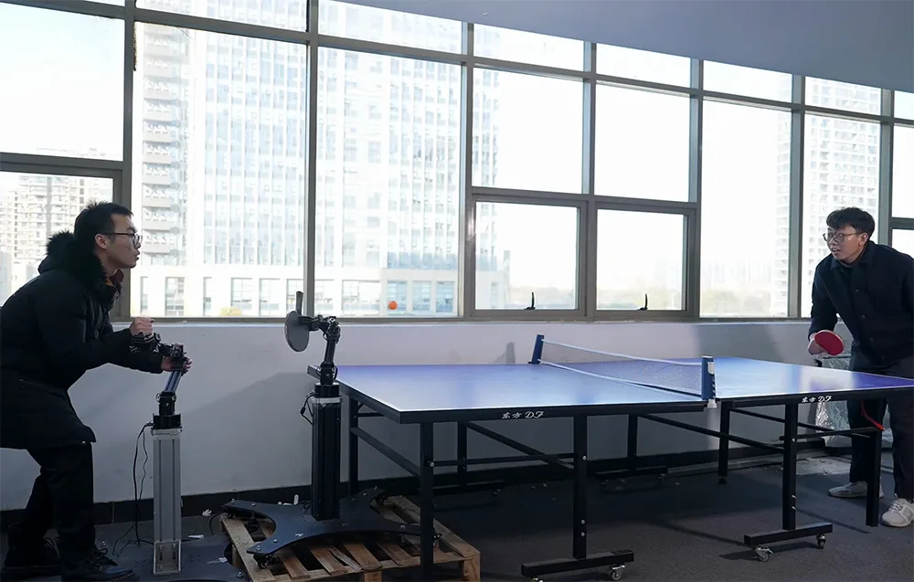 Man playing ping pong with a robotic arm controlled by another man