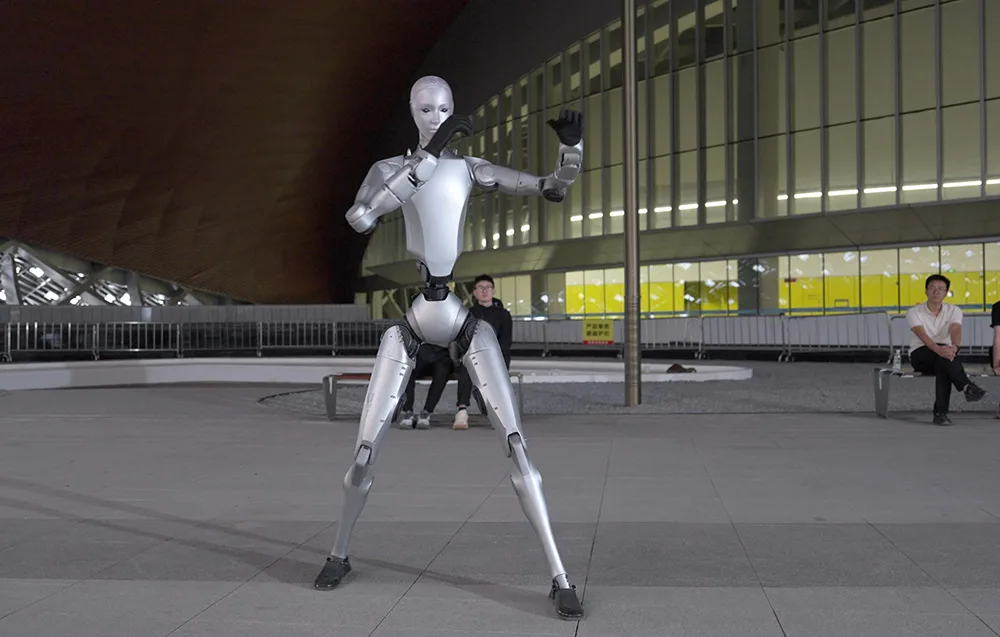UNITREE H2 humanoid robot in a defensive pose near modern architecture at night