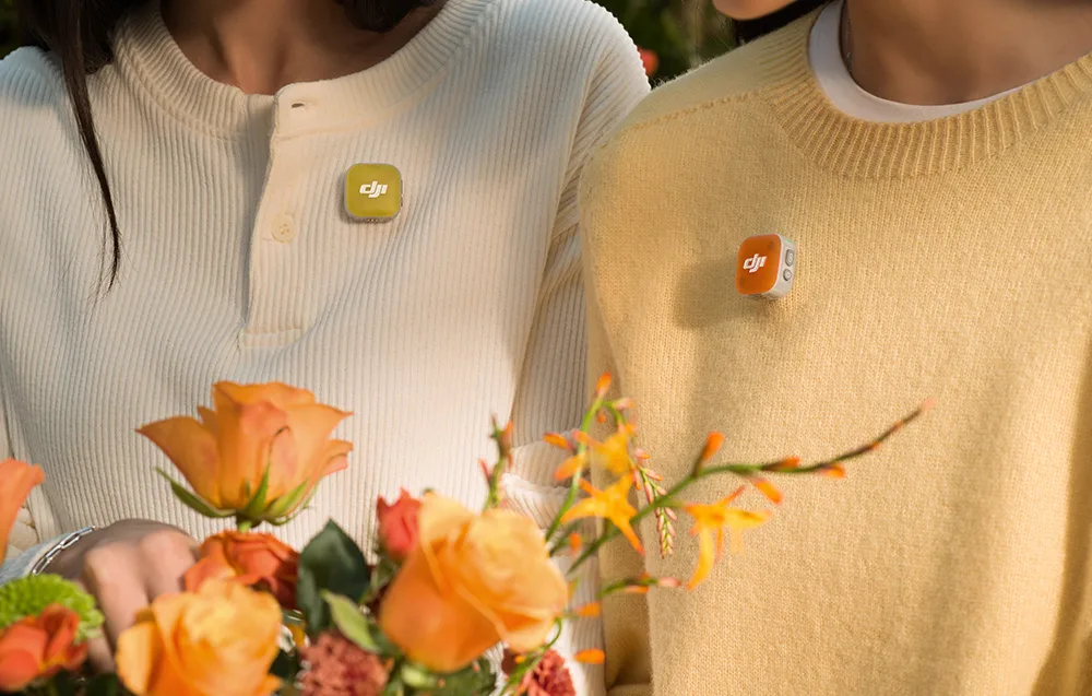 Two people in light sweaters wearing DJI microphones with yellow and orange magnetic covers, standing next to a bouquet of roses
