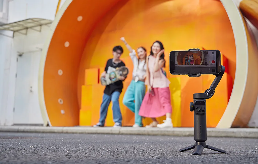 Gimbal ustawiony na mini statywie, nagrywający trójkę przyjaciół pozujących radośnie na pomarańczowej scenie.