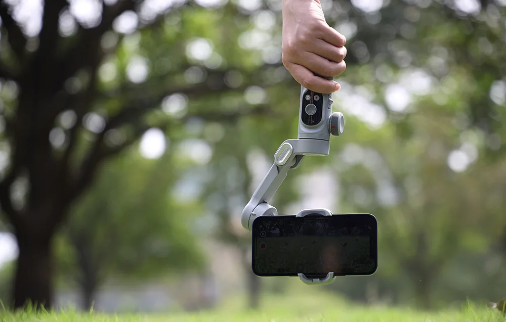 Dłoń trzyma gimbal ze smartfonem skierowanym w dół podczas nagrywania w plenerze.