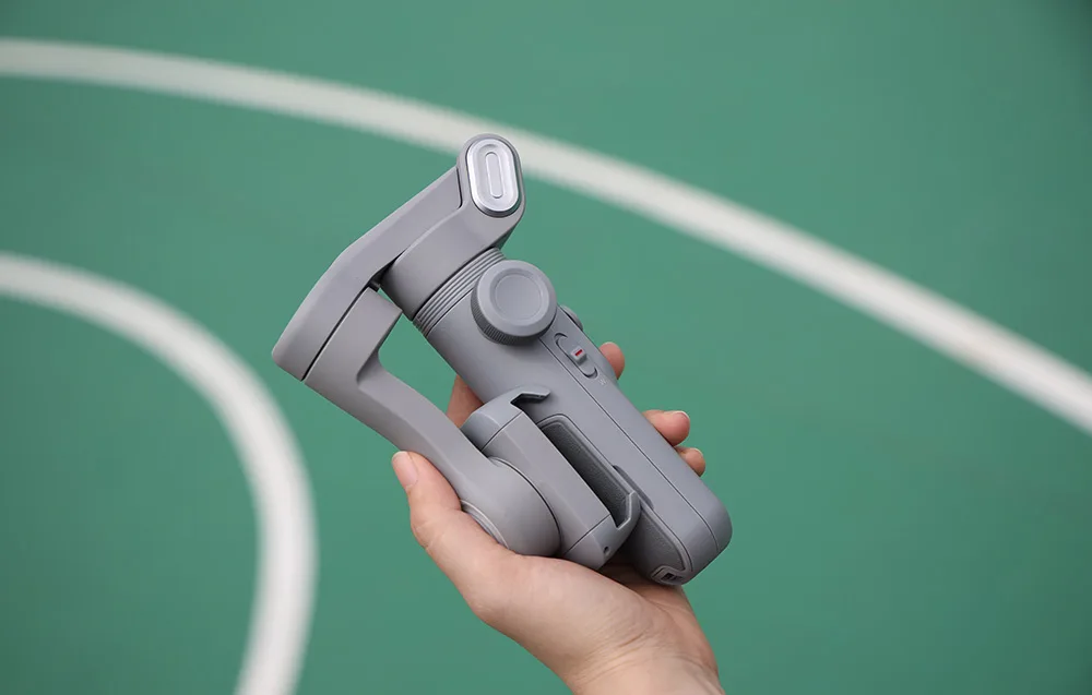 Dłoń trzyma złożony, kompaktowy gimbal na tle zielonego boiska sportowego.