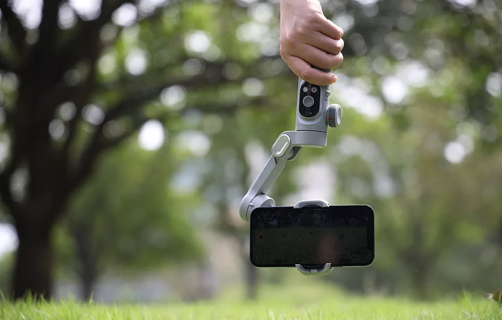 Gimbal ze smartfonem trzymany nad trawą podczas nagrywania w plenerze.