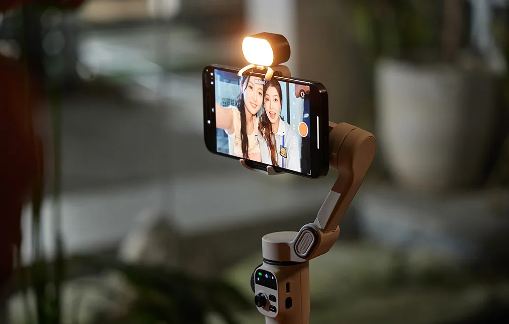 Smartfon zamocowany w beżowym gimbalu z włączoną lampą, wyświetlający selfie dwóch kobiet.
