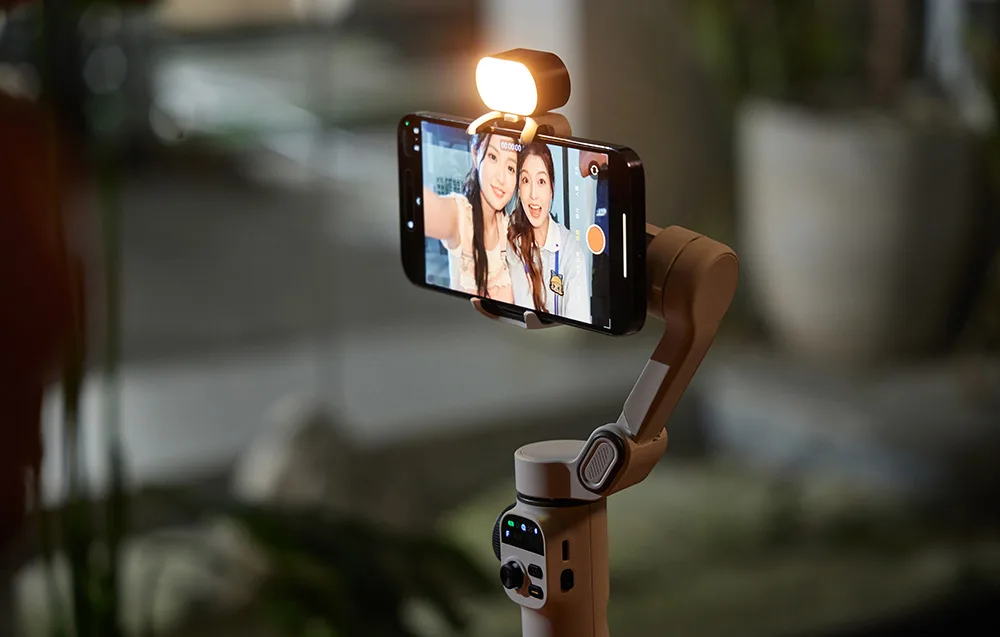 Smartfon zamocowany w beżowym gimbalu z włączoną lampą, wyświetlający selfie dwóch kobiet.
