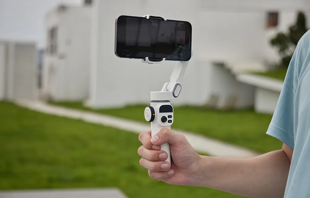 Dłoń trzymająca biały gimbal ze smartfonem w poziomie podczas nagrywania na świeżym powietrzu.