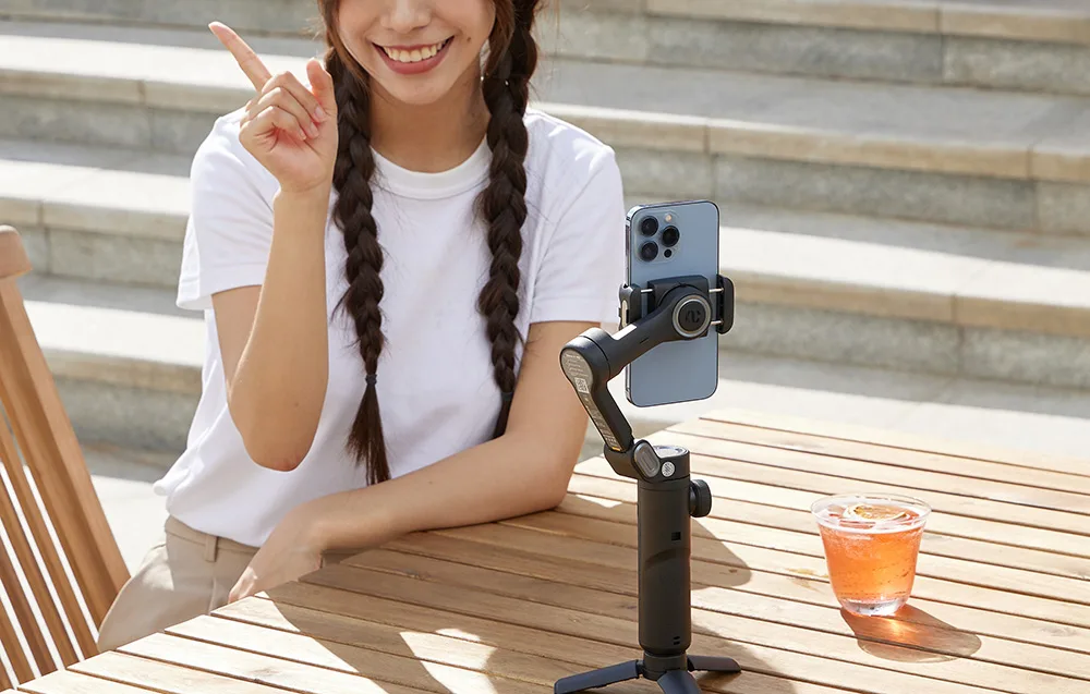 Woman pointing while recording with a smartphone mounted on the AOCHUAN XE gimbal on a tripod at a table