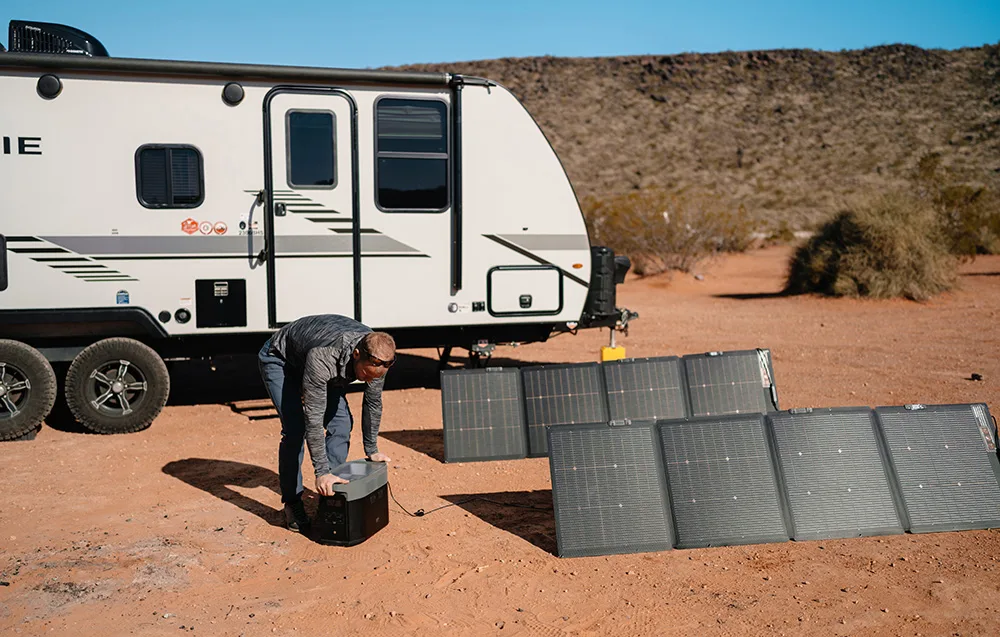 Man placing a portable power station on the ground next to unfolded solar panels and a camper trailer