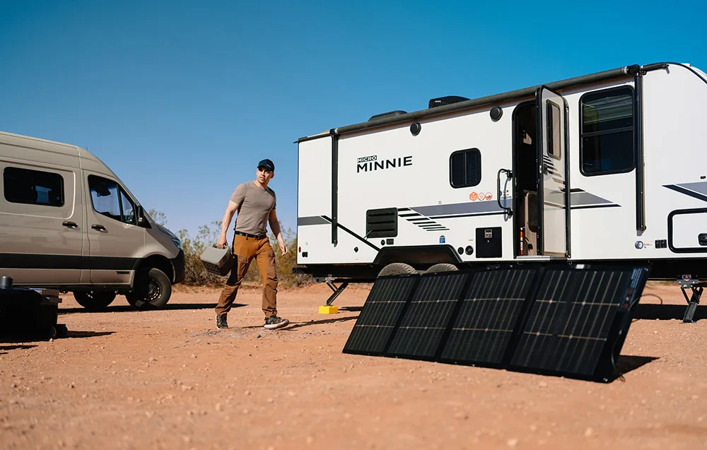 Man walking with a portable power station across sandy terrain next to a white camper trailer