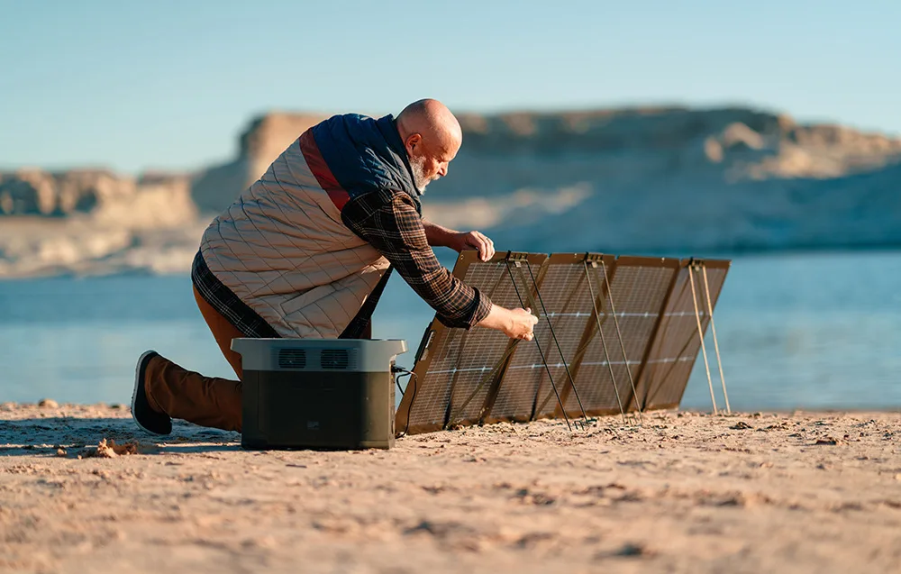 Man kneeling on a sandy shore while setting up the kickstands of a portable solar panel