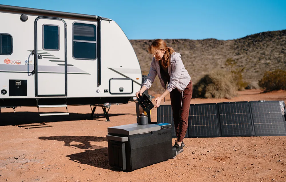 Woman using a portable camping fridge placed on the ground next to a camper trailer