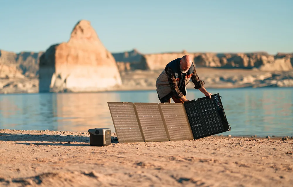Man unfolding a portable solar panel on a sandy shore near the water with a power station beside it