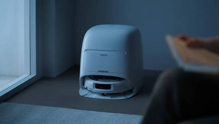 DJI Romo robot vacuum in the docking station in a dim room next to a person reading a book