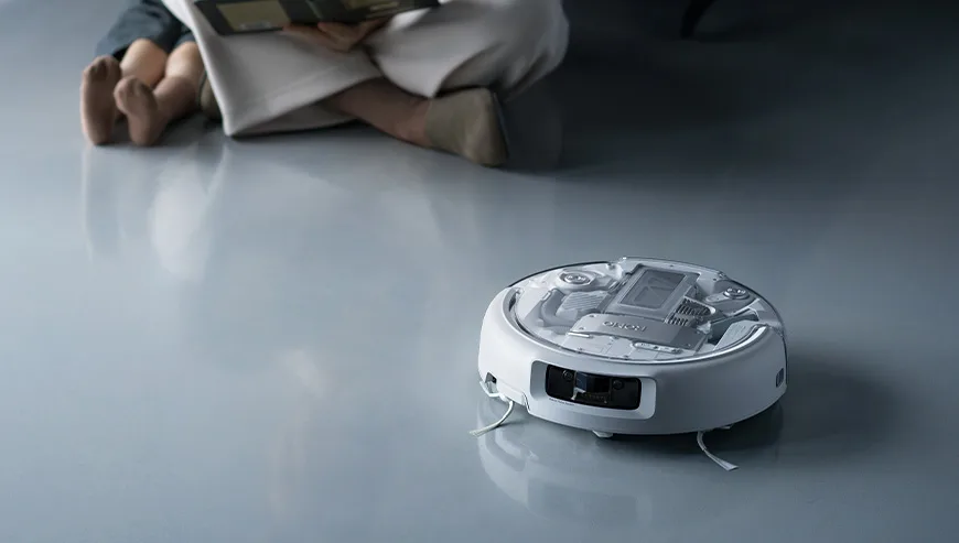 DJI Romo robot vacuum cleaning the floor while a person reads a book in the background