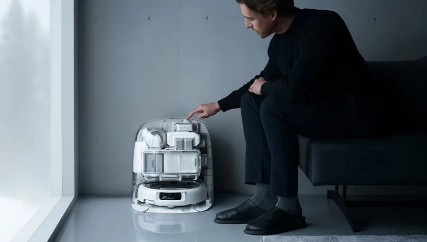 Man sitting on a sofa touching the top of the DJI ROMO docking station with the robot placed inside