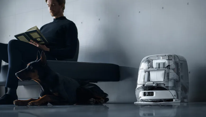 Man sitting on a sofa reading a book next to a dog and the DJI ROMO docking station