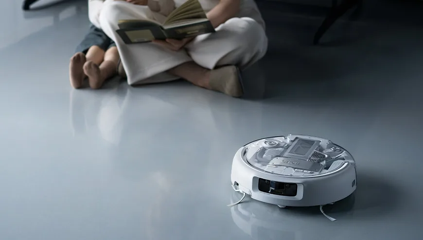 DJI ROMO robot vacuum moving across the floor near a person sitting and reading a book