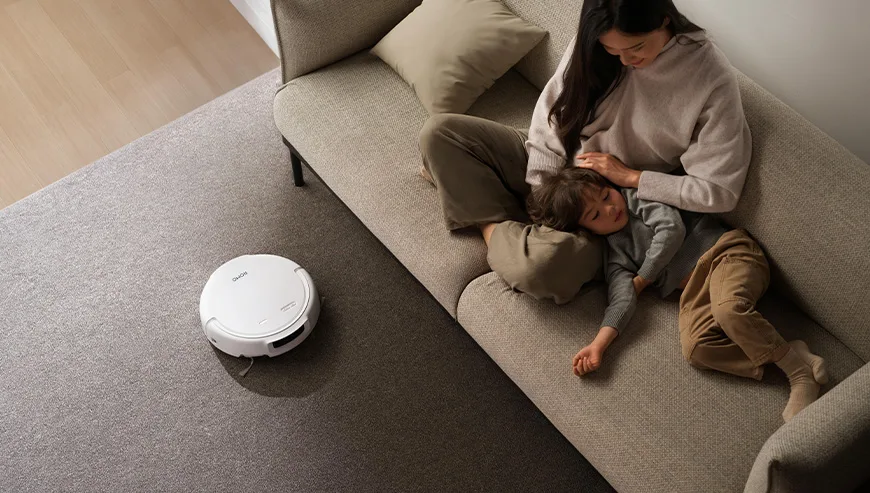 Kobieta siedzi na kanapie z dzieckiem, a robot sprzątający ROMO porusza się po dywanie